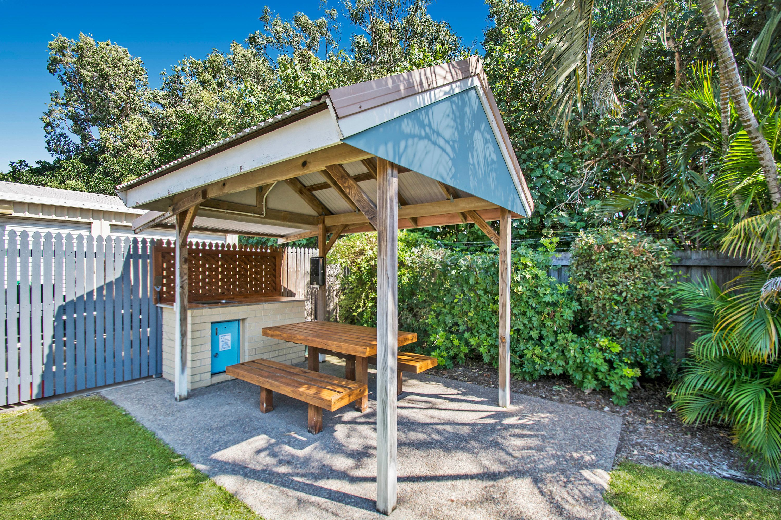 A covered outdoor seating area with wooden benches and a blue door, surrounded by lush greenery and a wooden fence.