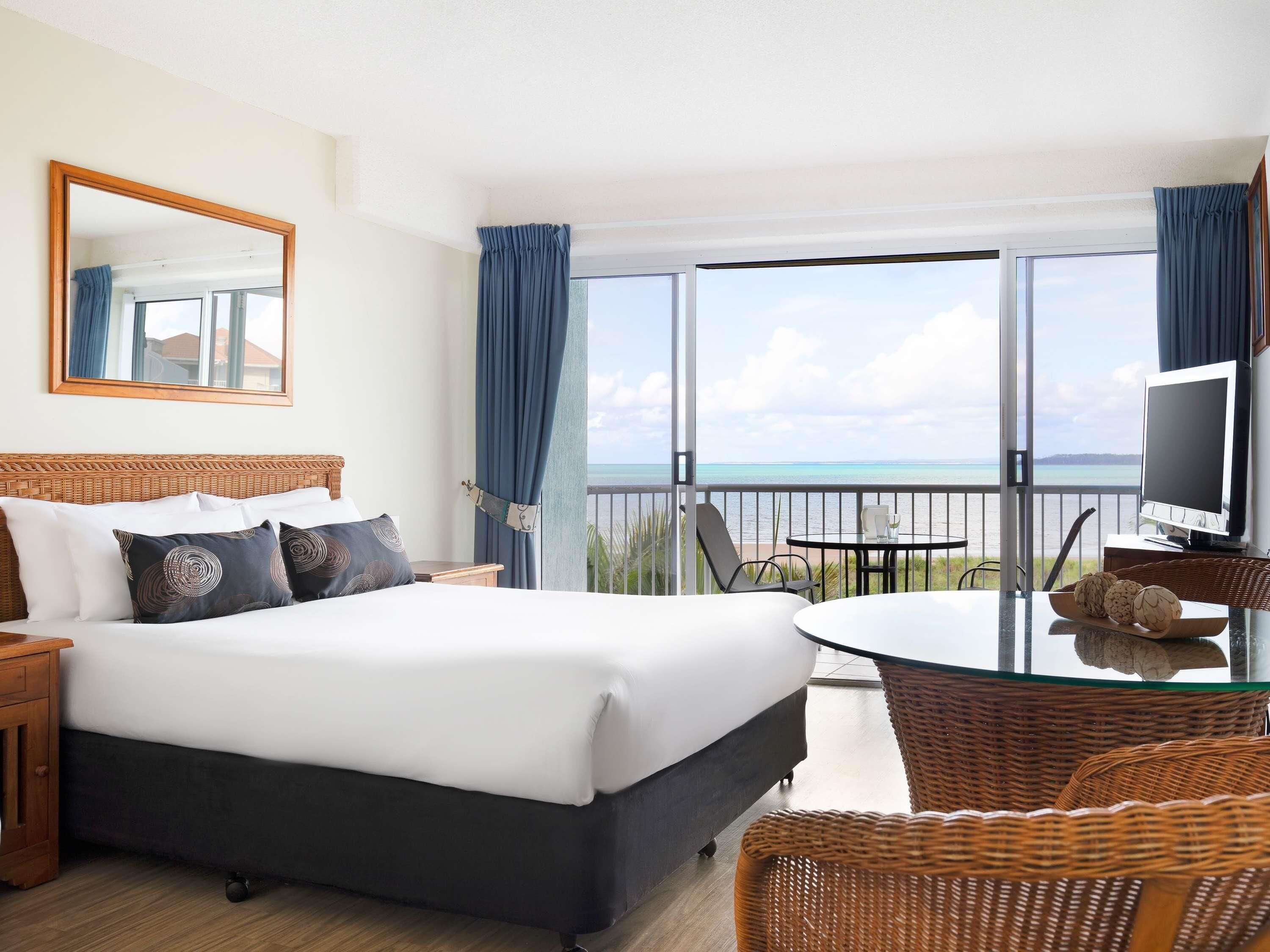 A hotel room with a bed, a TV, a balcony with a view of the sea, and wicker furniture.