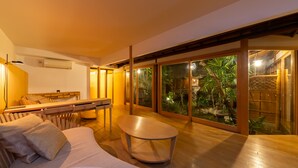 Una sala de estar moderna con un sofá, una mesa redonda de centro y un comedor con una mesa y sillas de madera. Amplias puertas correderas de vidrio dan a vistas de un jardín tropical.