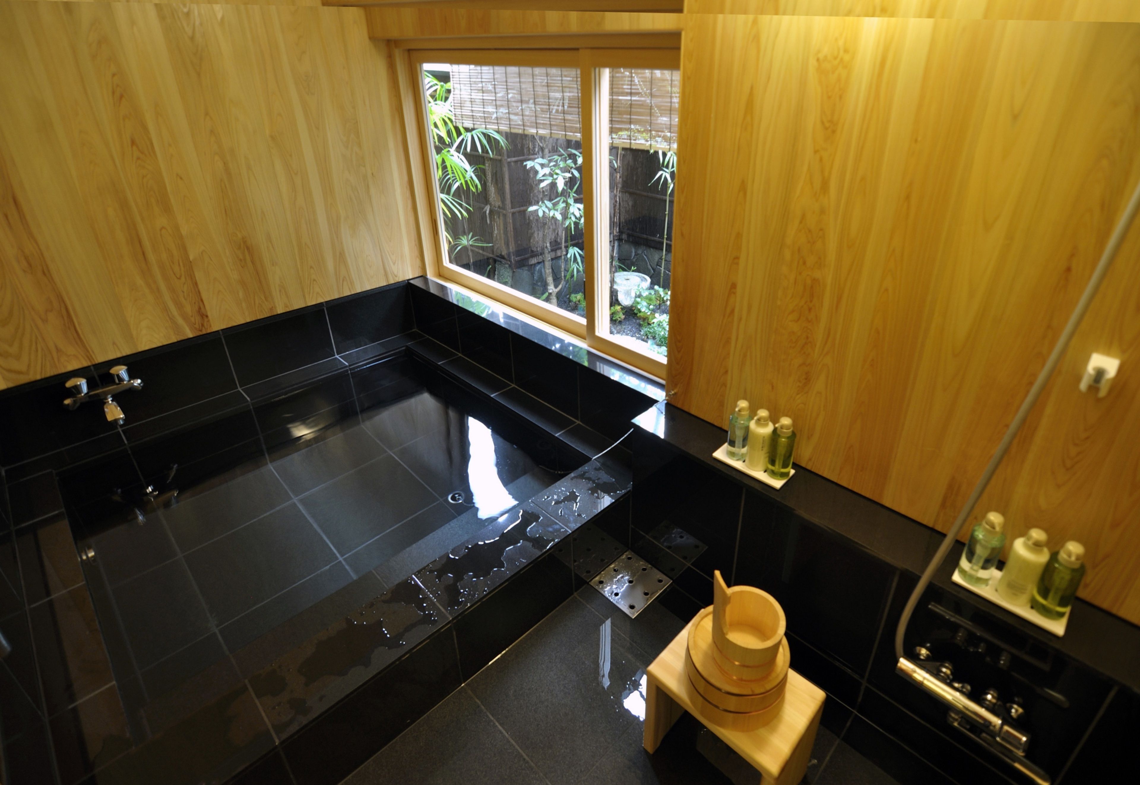 A modern bathroom with a black bathtub, wooden cabinets, and a window with a view of greenery.
