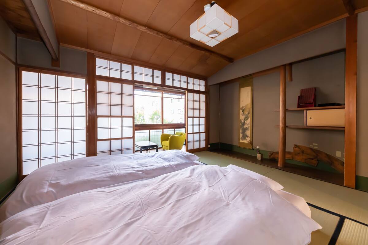 A traditional Japanese-style room with tatami flooring, a large bed, a small table with a chair, and sliding doors.