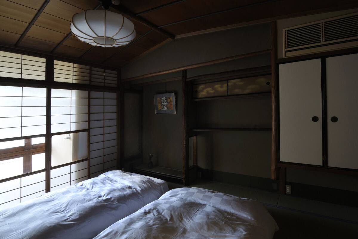 A traditional Japanese room with tatami flooring, a low wooden ceiling, and sliding doors. There are two beds with white bedding and a hanging lamp.