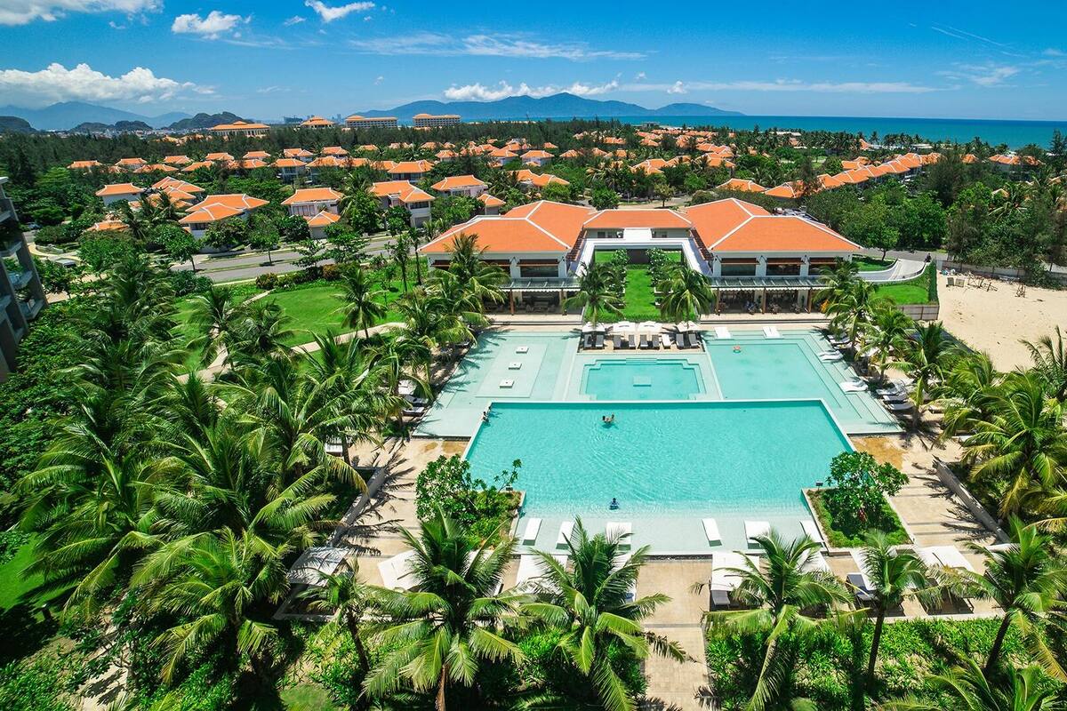 A resort with a central swimming pool, surrounded by palm trees and a view of the ocean.