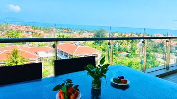 Un balcón con vista a una zona residencial, una mesa con una planta y fruta, y una barandilla de vidrio.