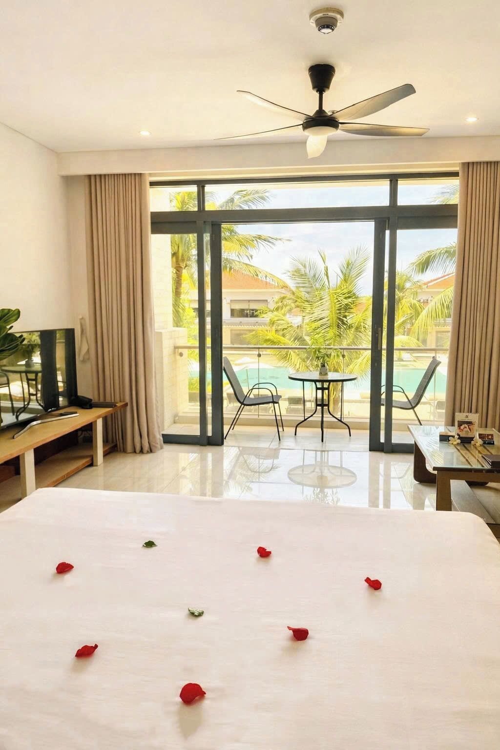 A hotel room with a bed, a desk, a chair, a table, a ceiling fan, and a large window with a view of palm trees and a pool.