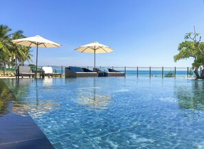 Une piscine d’un bleu éclatant, avec des chaises longues et des parasols, un palmier, et une vue sur l’océan.