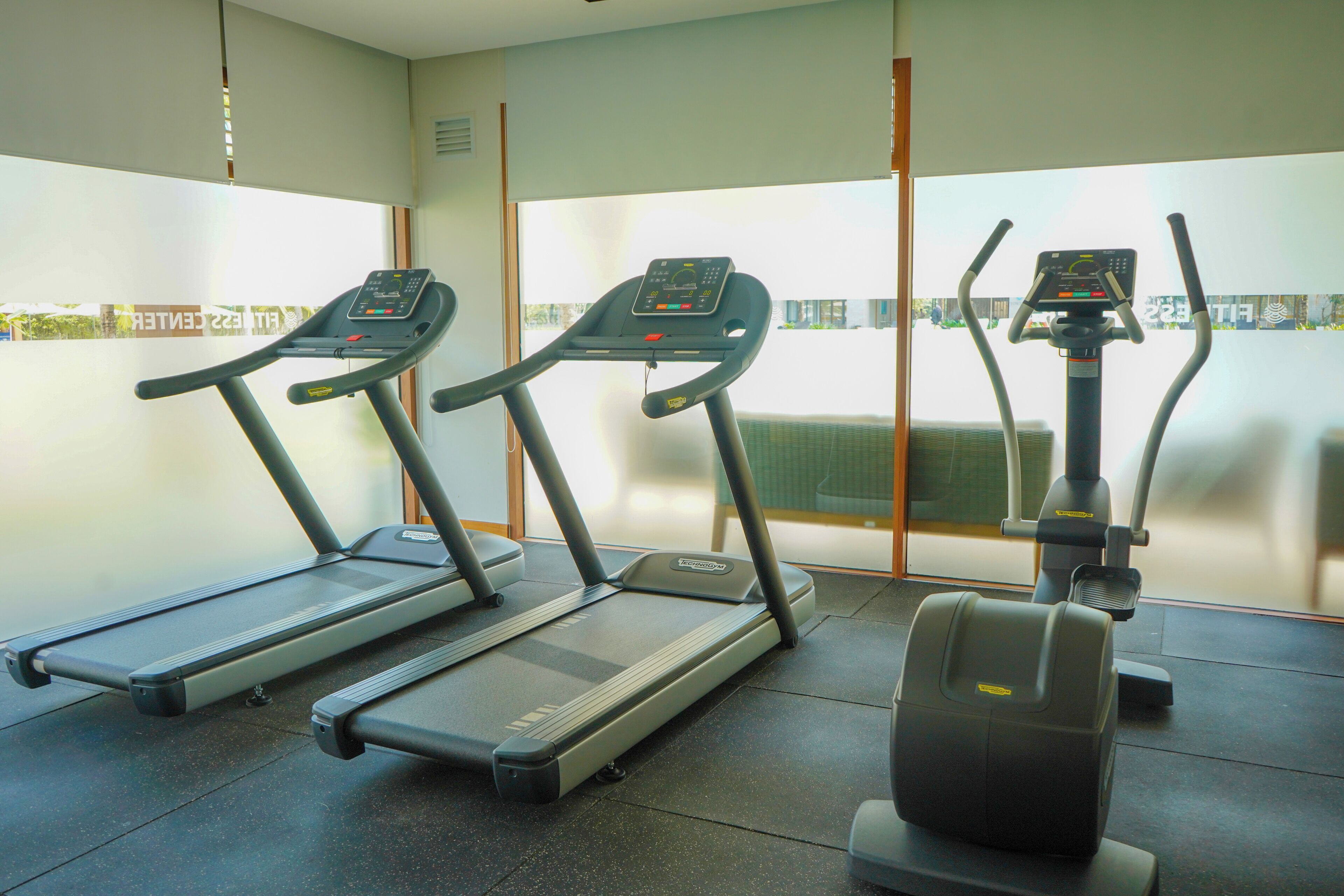Tres máquinas de ejercicio en un gimnasio con un amplio ventanal al fondo.