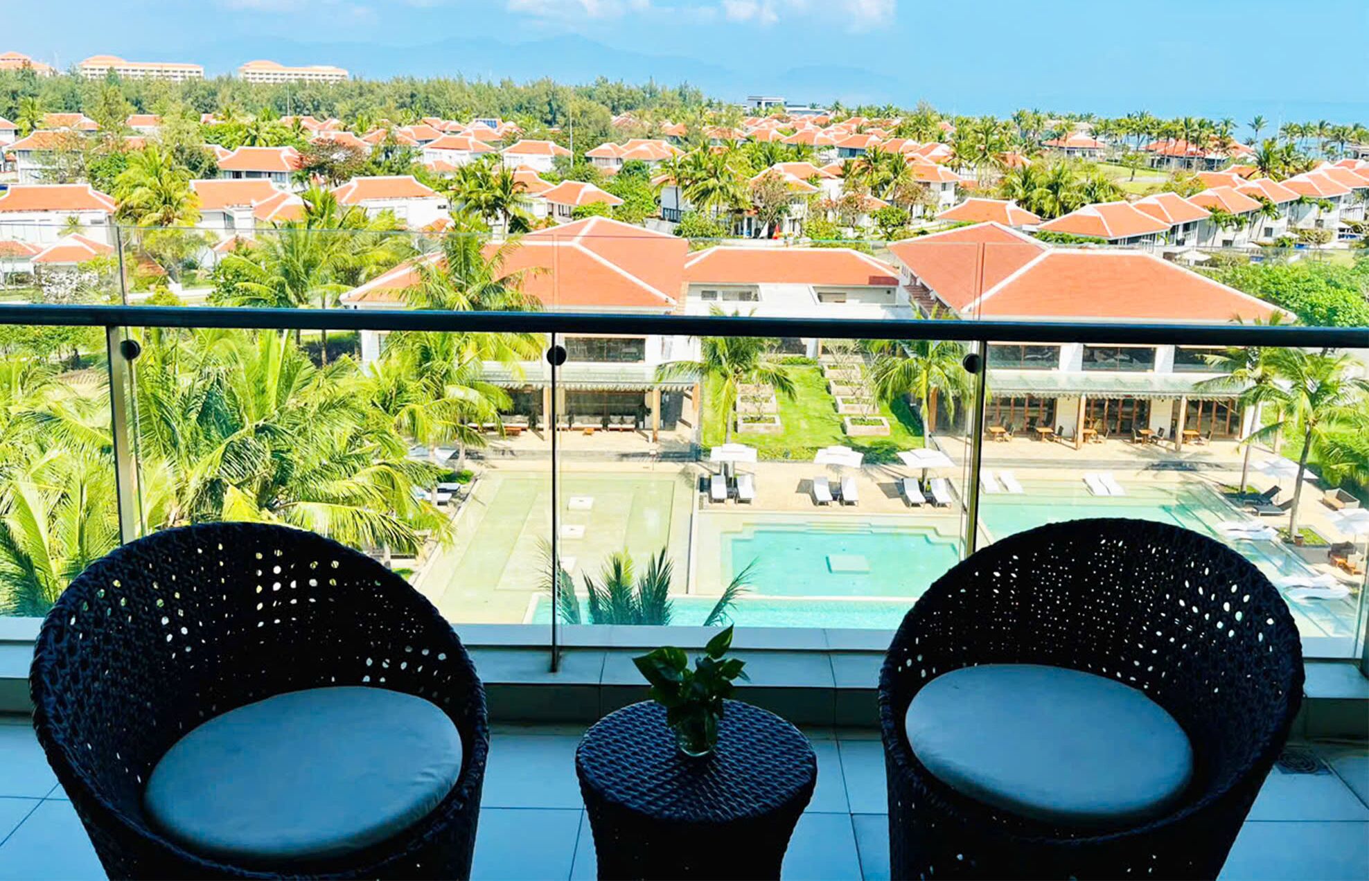 Un balcon avec deux fauteuils en osier, une vue sur un complexe hôtelier comprenant plusieurs bâtiments, une piscine et des palmiers.