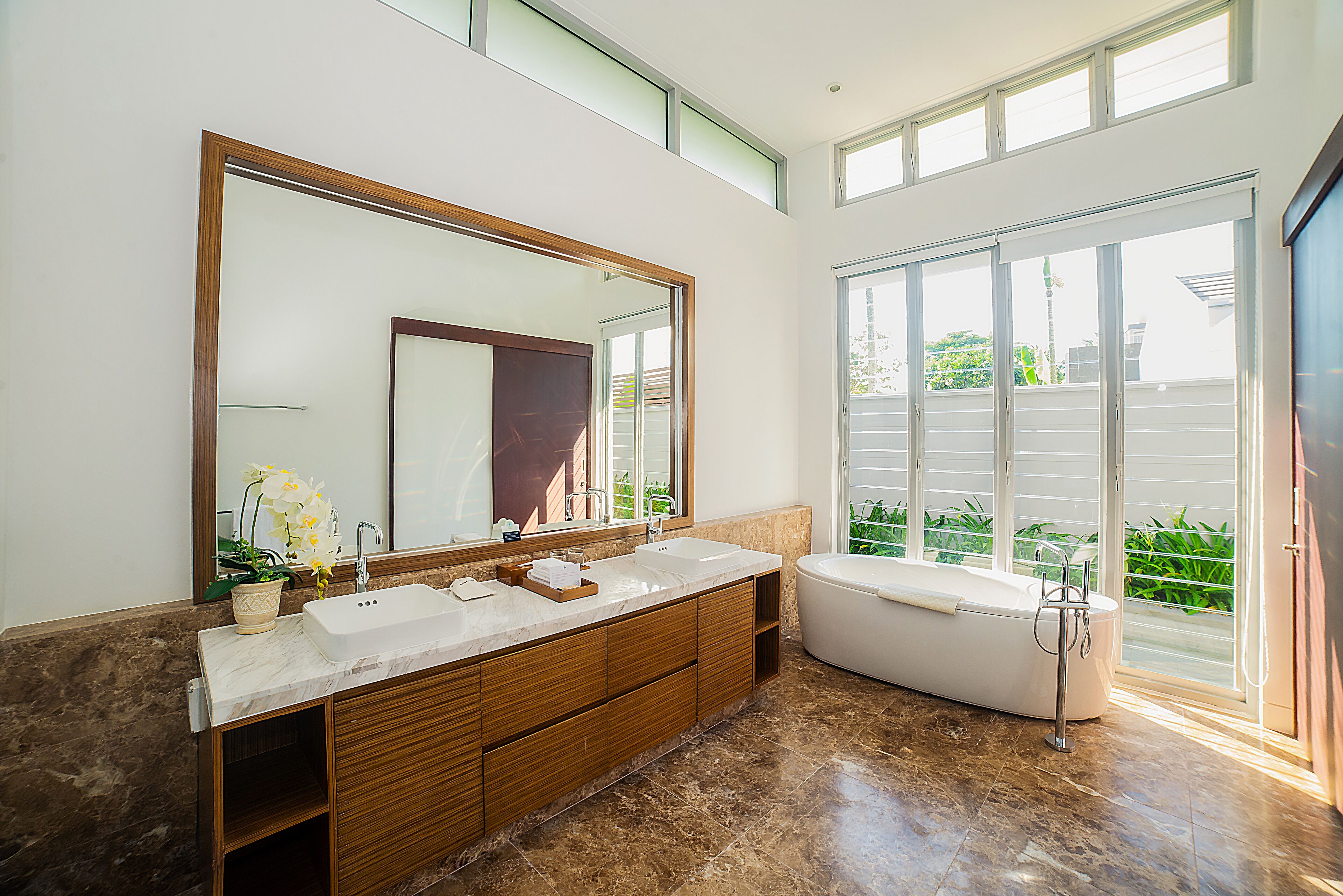 A modern bathroom with a large mirror, two sinks, a bathtub, and a view of greenery outside.