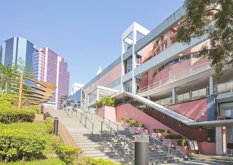 A modern building with a staircase leading to an upper level, surrounded by greenery and other buildings.