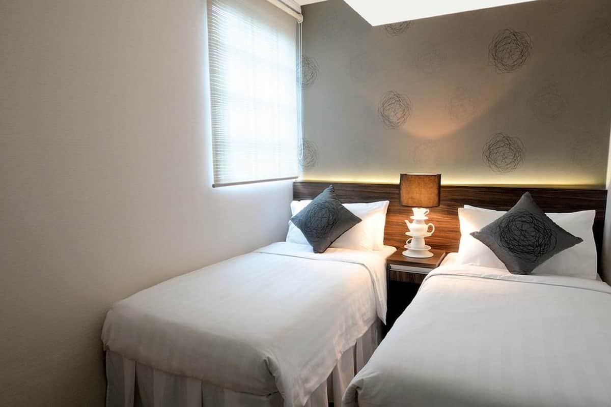 Ein Hotelzimmer mit zwei Einzelbetten, einem Nachttisch mit Lampe und einem Fenster mit Jalousien.