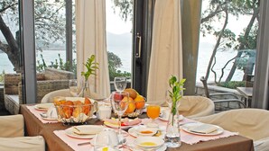Una mesa de desayuno dispuesta con platos, tazones, vasos y una cesta de fruta, con vistas a un paisaje pintoresco.