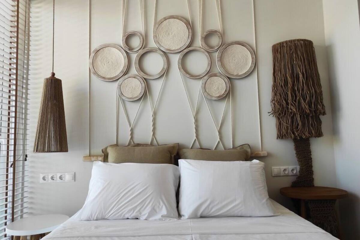 A bedroom with a bed, white pillows, a wall-mounted macrame headboard, and a woven lamp.