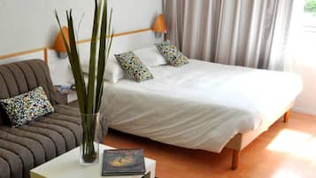 Een slaapkamer met een bed, een nachtkastje met een boek, een vaas met planten, een bank met gekleurde kussens en een raam met gordijnen.