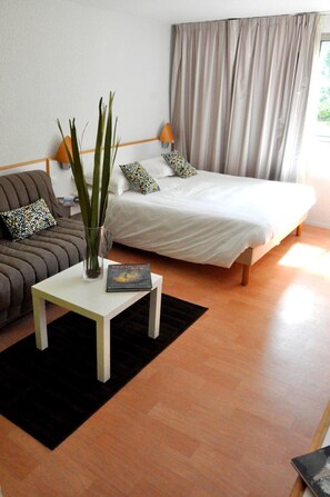 A bedroom with a bed, a bedside table with a book, a vase with plants, a sofa with patterned cushions, and a window with curtains.