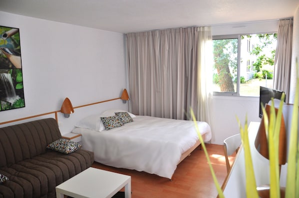 A bedroom with a bed, a sofa, a bedside table, a window with curtains, and a view of greenery outside.