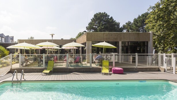 Una piscina con sillas de descanso y sombrillas, rodeada por una cerca blanca y un edificio con grandes ventanales.