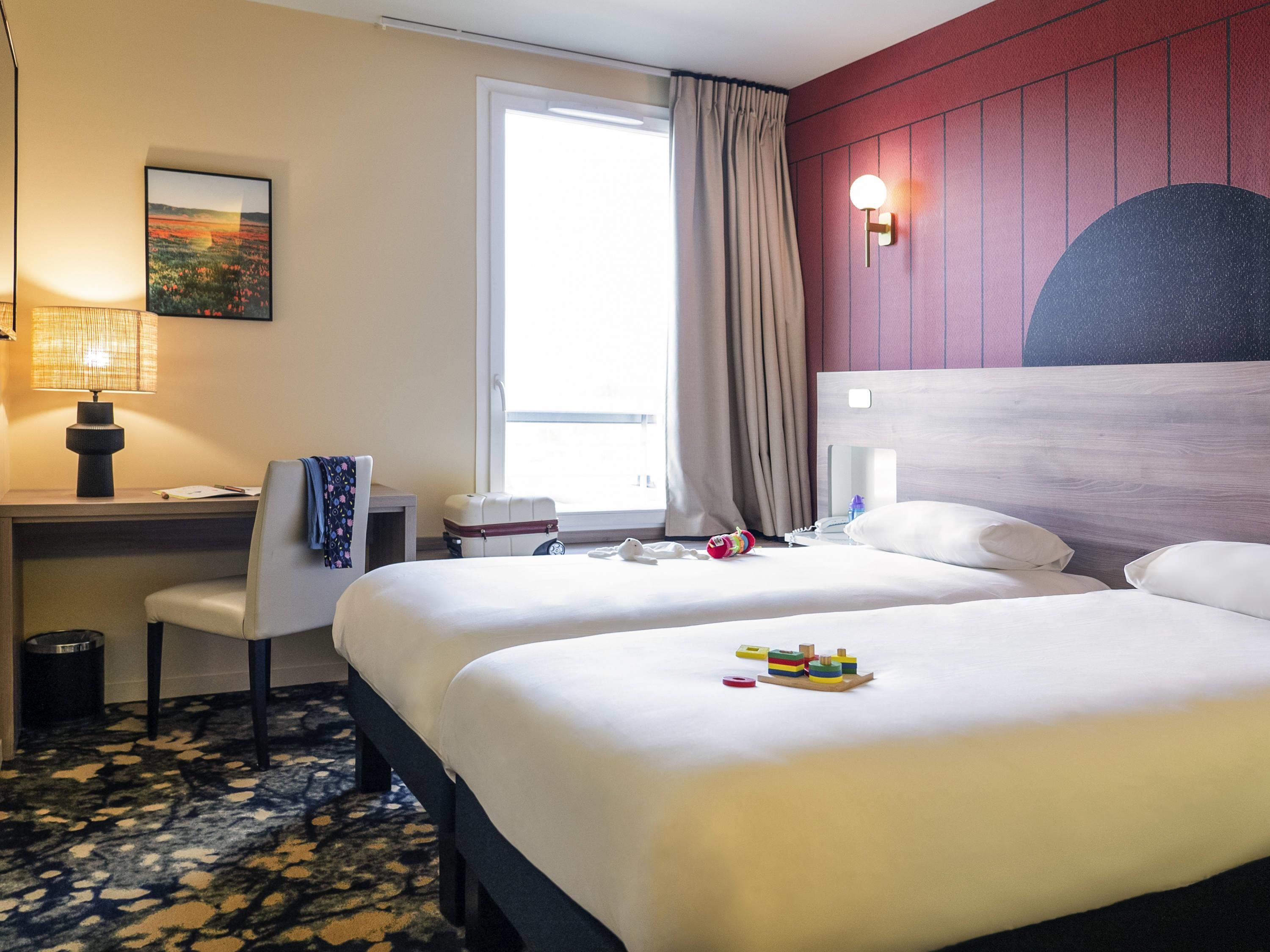 A hotel room with two beds, a desk, a chair, a lamp, a window with curtains, and a wall-mounted light fixture.