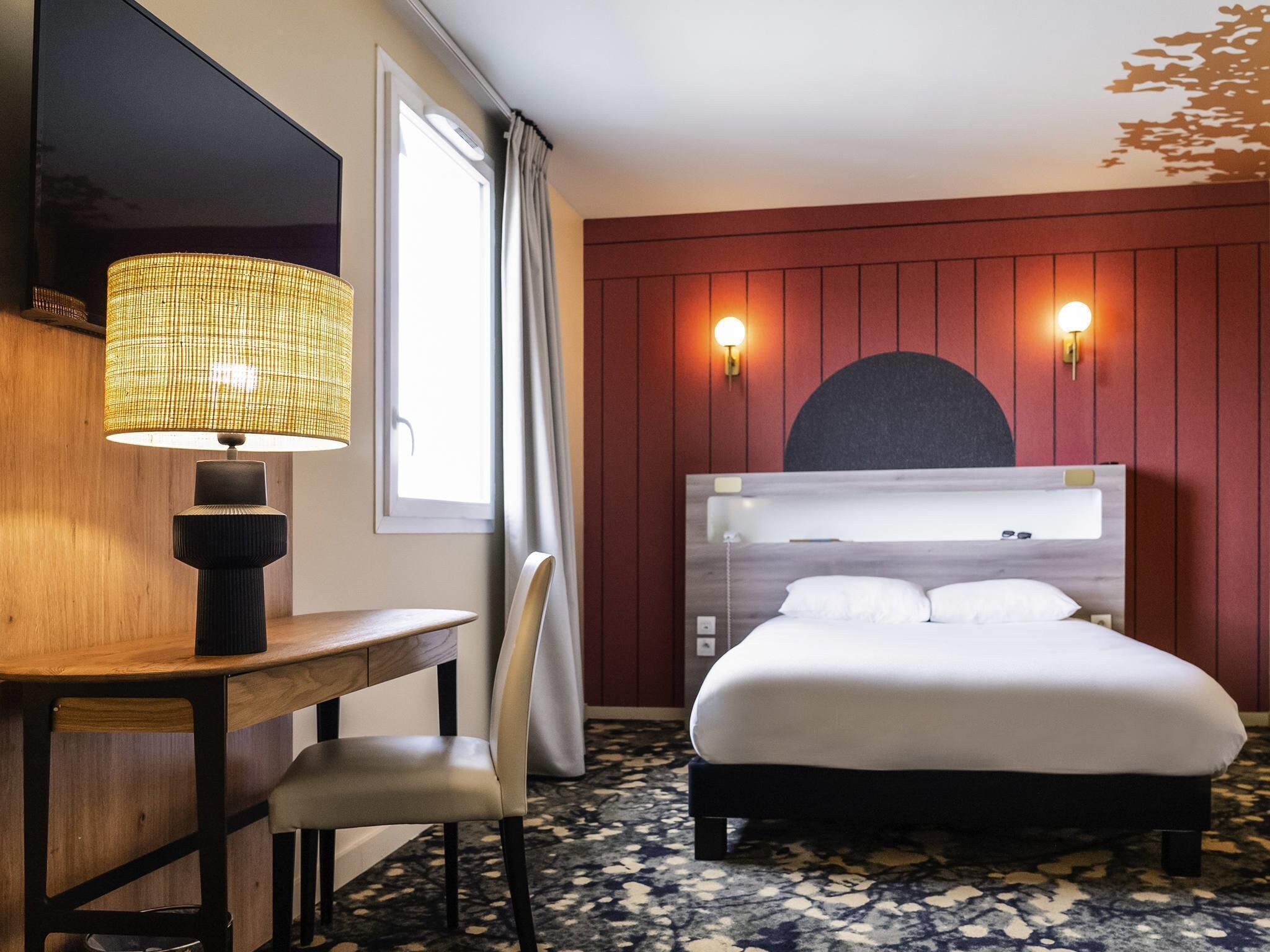 A hotel room with a bed, a desk, a chair, a lamp, and a window with curtains.