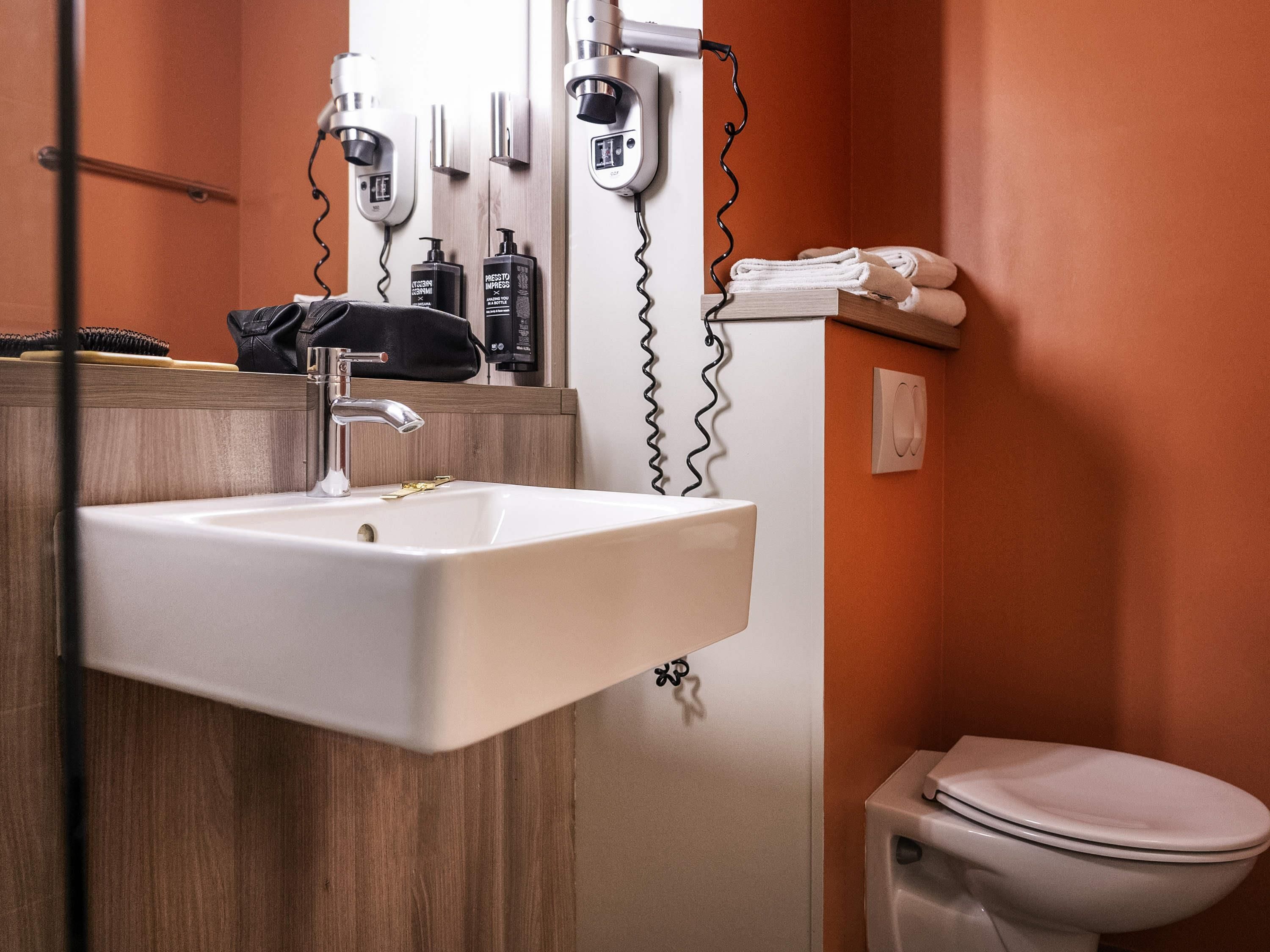 Un baño con un lavabo blanco, un espejo y un inodoro. Hay secadores de pelo y un perchero para toallas en la pared.