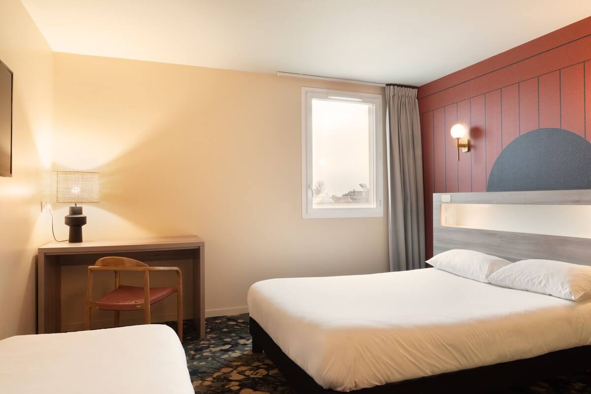 Een hotelkamer met twee bedden, een bureau met stoel, een lamp en een raam met gordijnen.