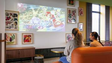 Ein Raum mit einem großen Bildschirm, der ein Videospiel zeigt, Sitzbereichen und verschiedenen gerahmten Kunstwerken an den Wänden.