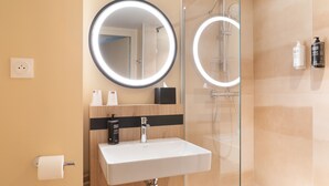 Une salle de bain moderne avec un lavabo blanc, deux miroirs éclairés, une douchette et une cabine de douche en verre.