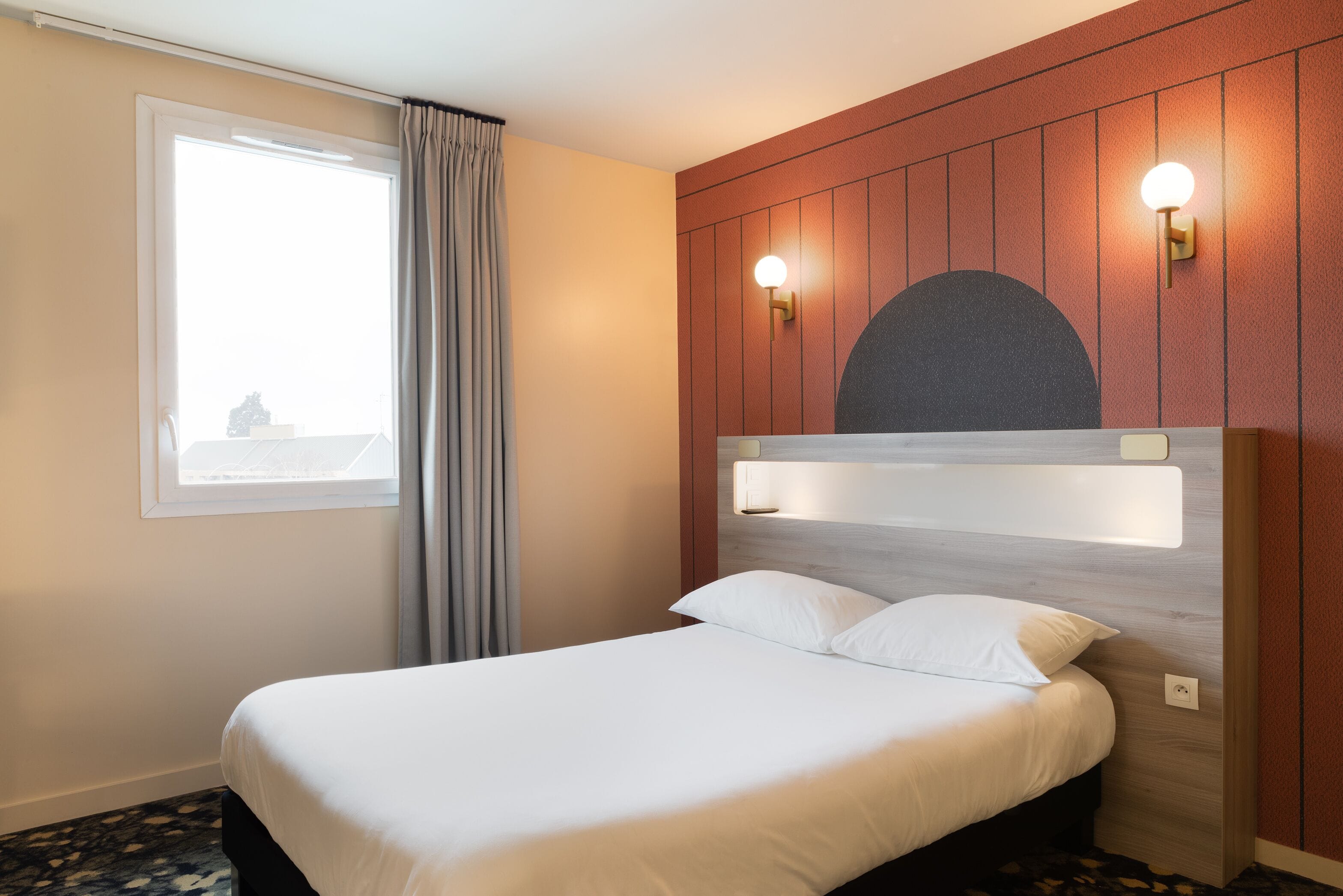 A hotel room with a bed, a window with curtains, and wall-mounted lighting.