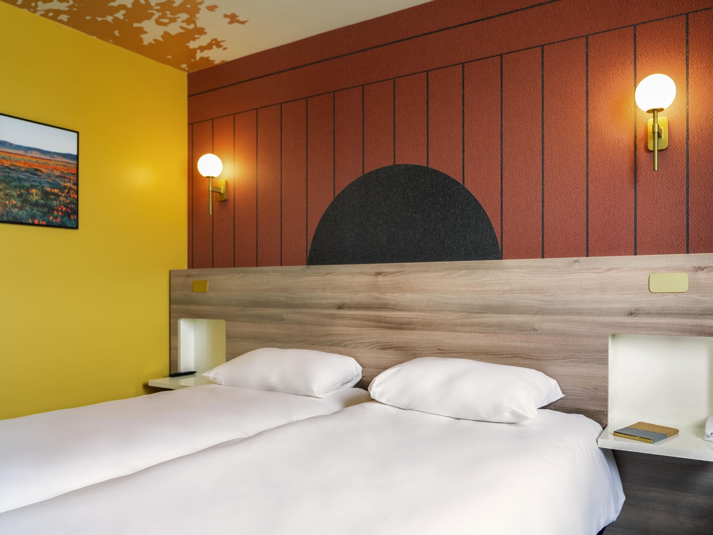 Habitación de hotel con cabecera de madera, dos mesitas de noche con lámparas, cama con ropa blanca y una obra de arte colgada en la pared.