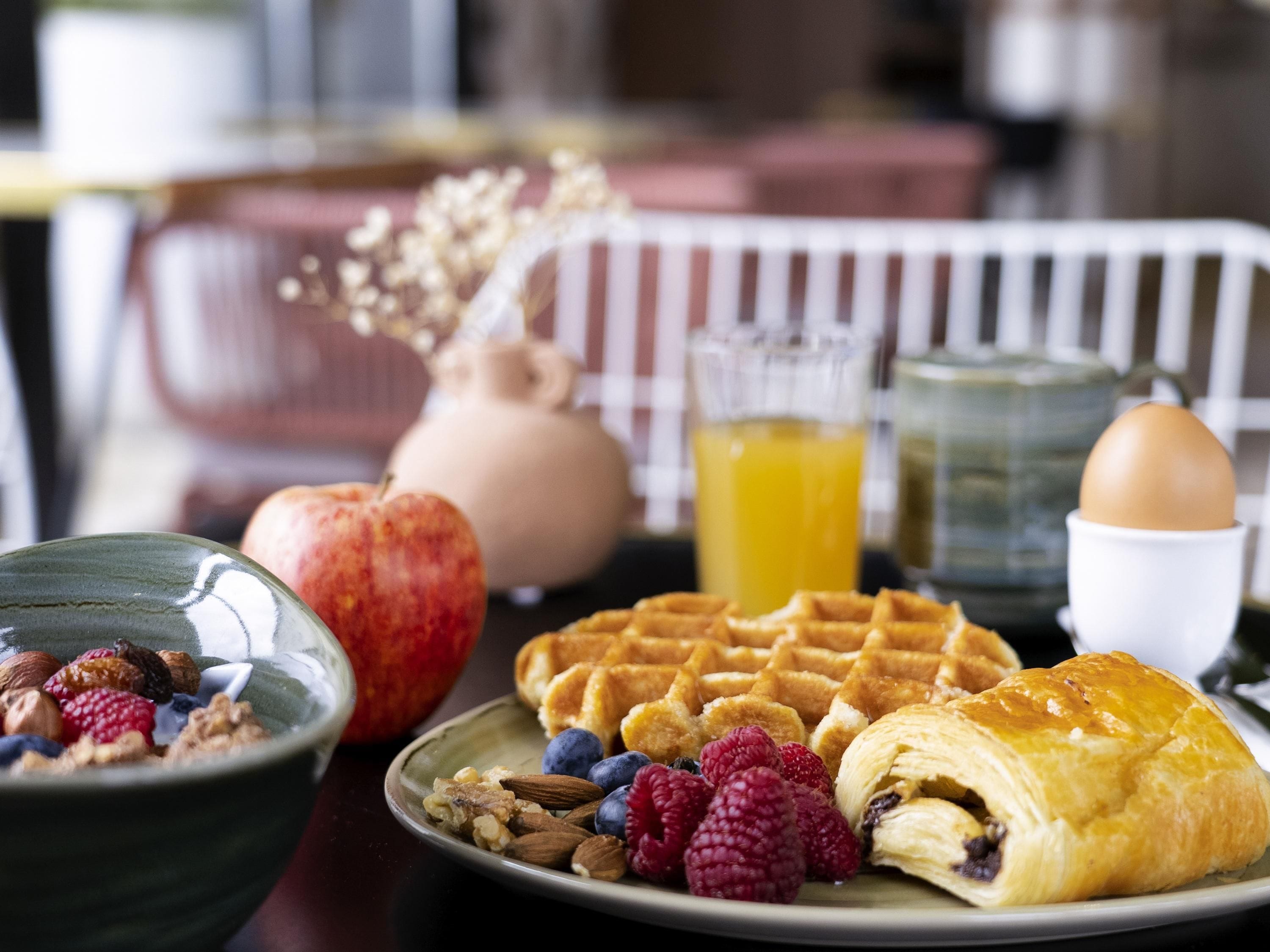 Una mesa de desayuno con waffles, un croissant, un vaso de jugo de naranja, un tazón de cereal y un plato con frutas y nueces.