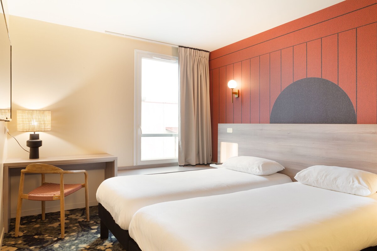 Une chambre d’hôtel avec deux lits, un bureau, une chaise, une lampe et une grande fenêtre avec des rideaux.