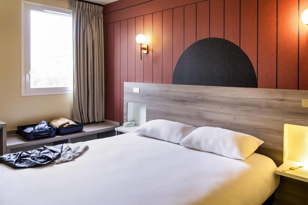 Hotelkamer met een bed, nachtkastje, raam met gordijnen, een wandlamp en een gedeeltelijk zicht op een roomdivider.