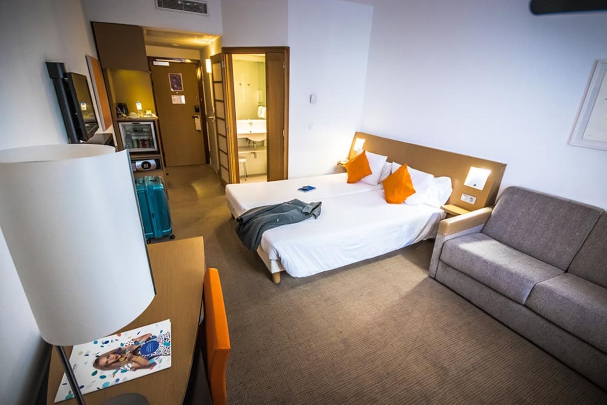 Habitación de hotel con cama, sofá, escritorio, televisor y baño visible a través de una puerta abierta.