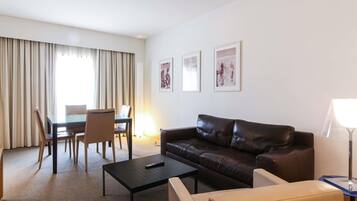A hotel room with a leather sofa, a small coffee table, a dining table with chairs, and a lamp.