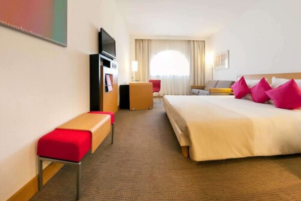 Una habitación de hotel con cama, escritorio, televisor y ventana con cortinas.