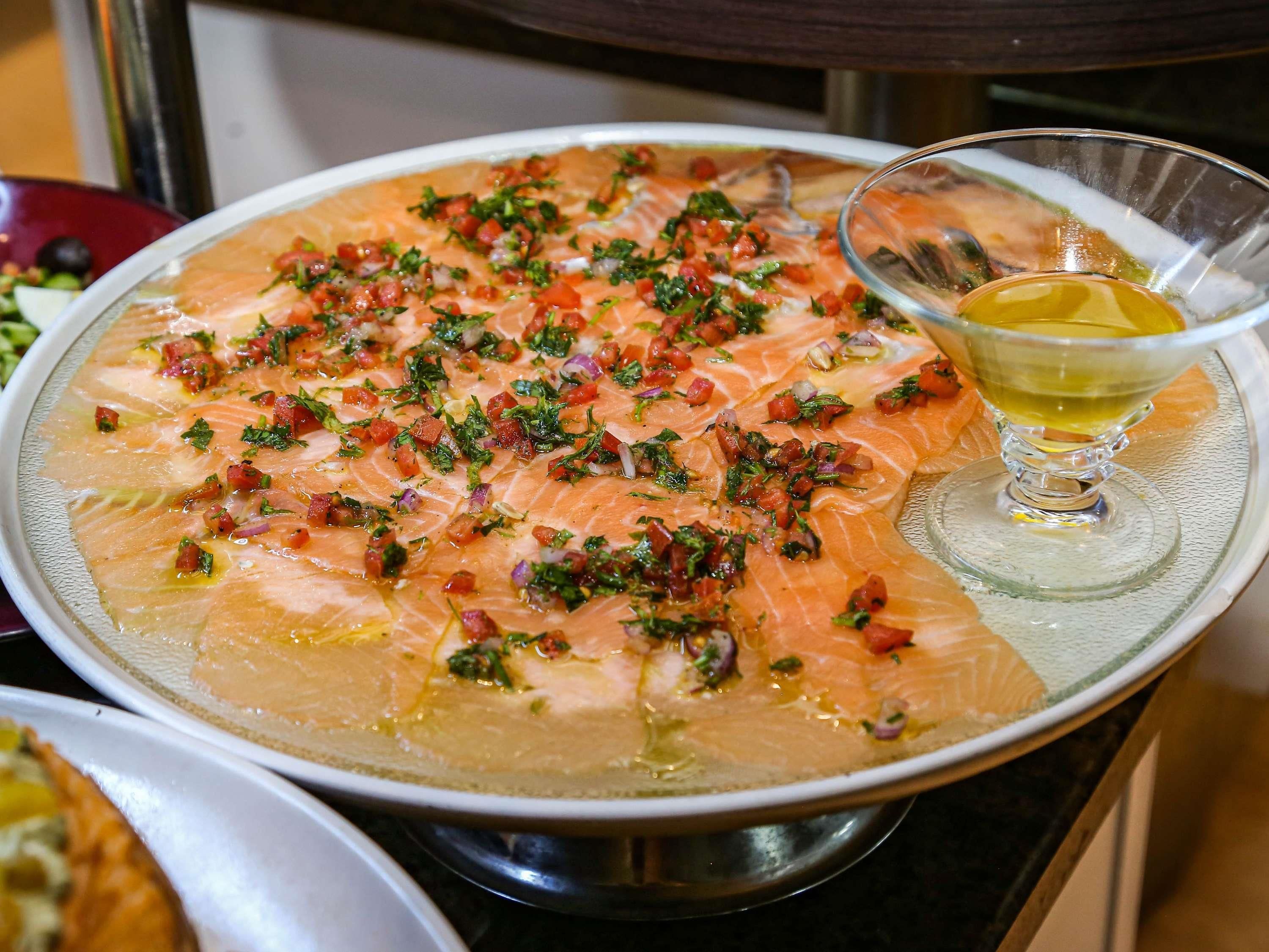 Un plato grande de salmón adornado con hierbas y tomates en cubos, acompañado de un vaso de aceite de oliva.