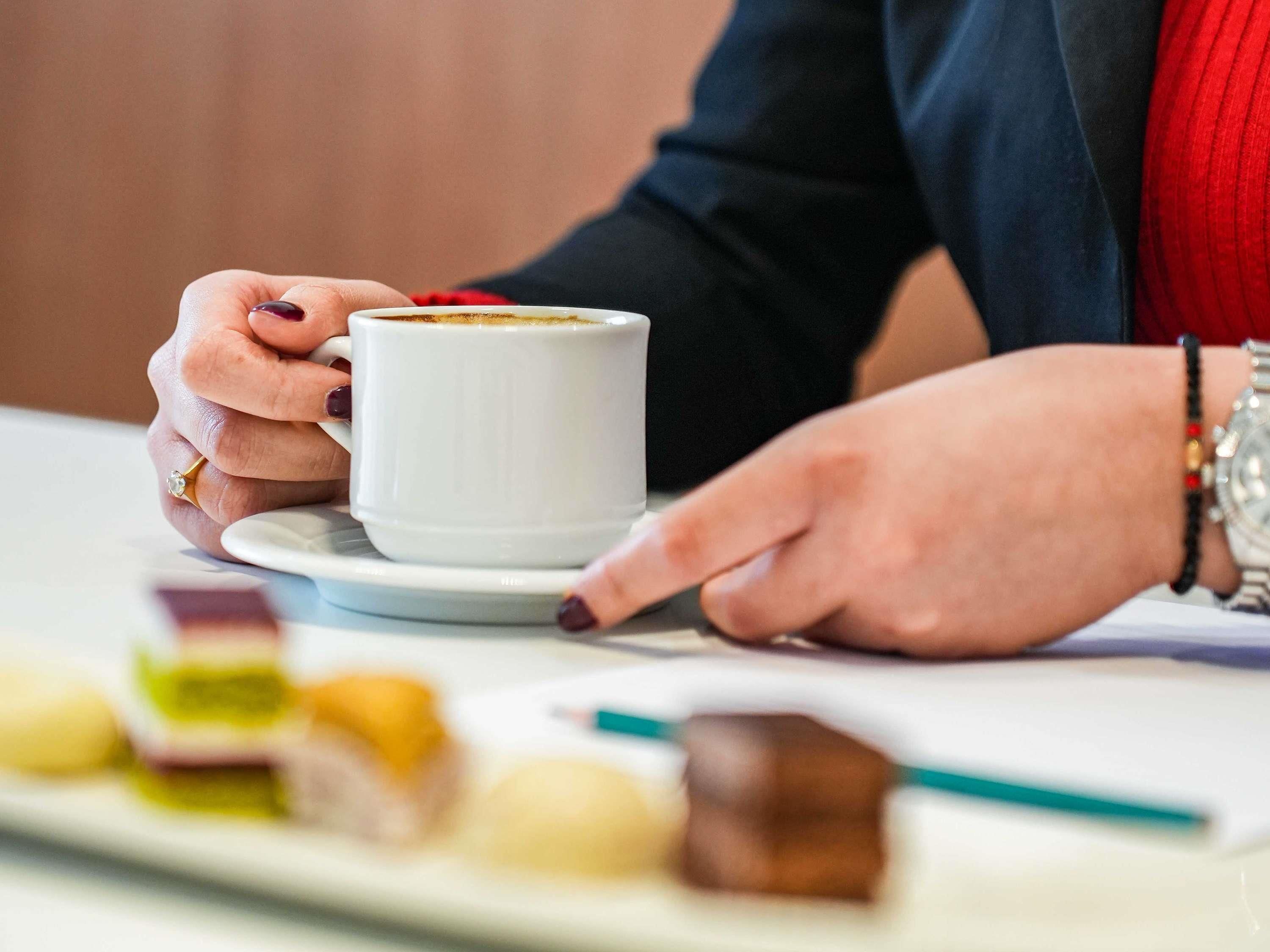Eine Person hält eine weiße Tasse mit einem Getränk und sitzt an einem Tisch mit einer Speisenplatte.