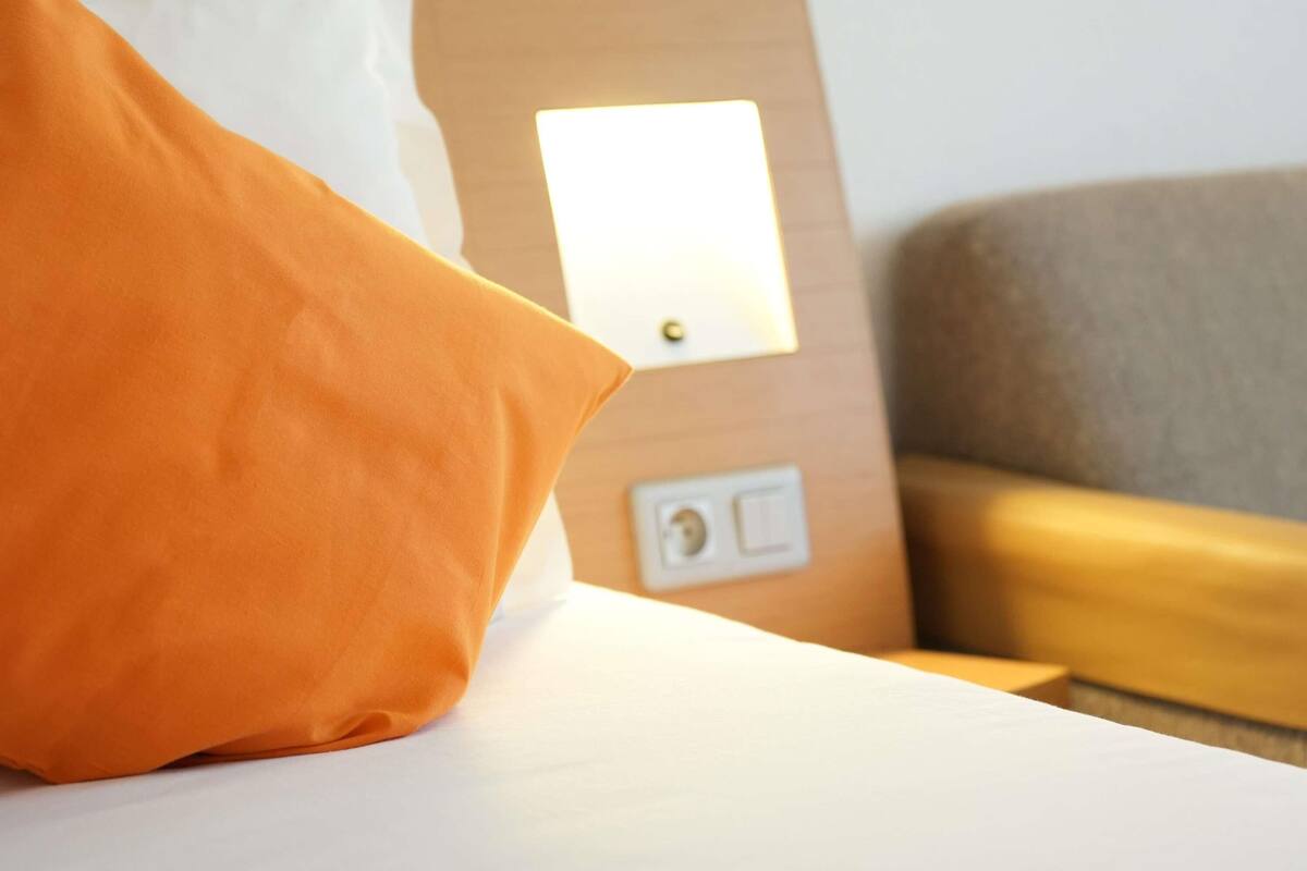 A bed with an orange pillow, a bedside table, and a nightstand with a lamp.