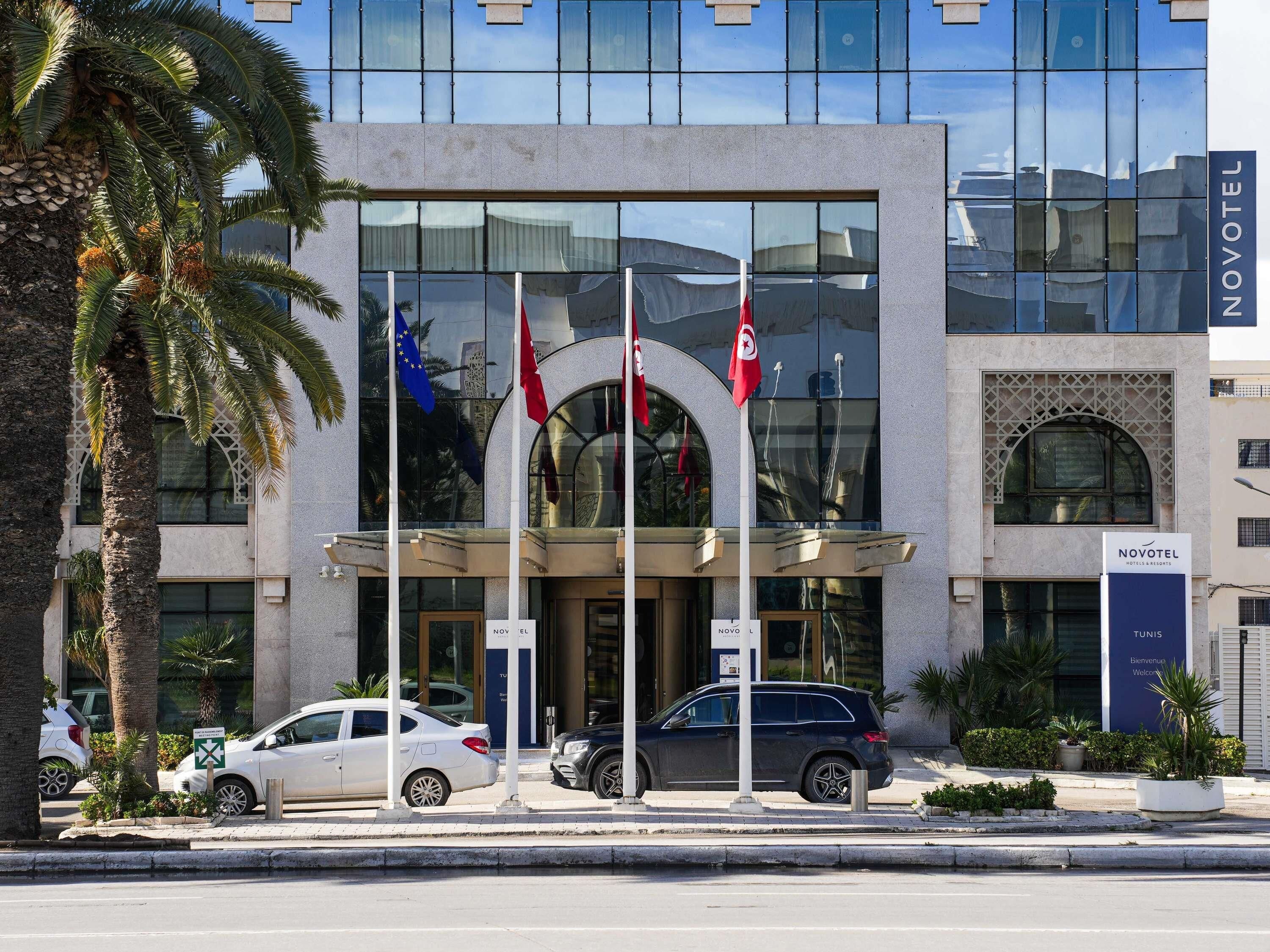 A hotel entrance with flags, a modern facade, and parked cars.