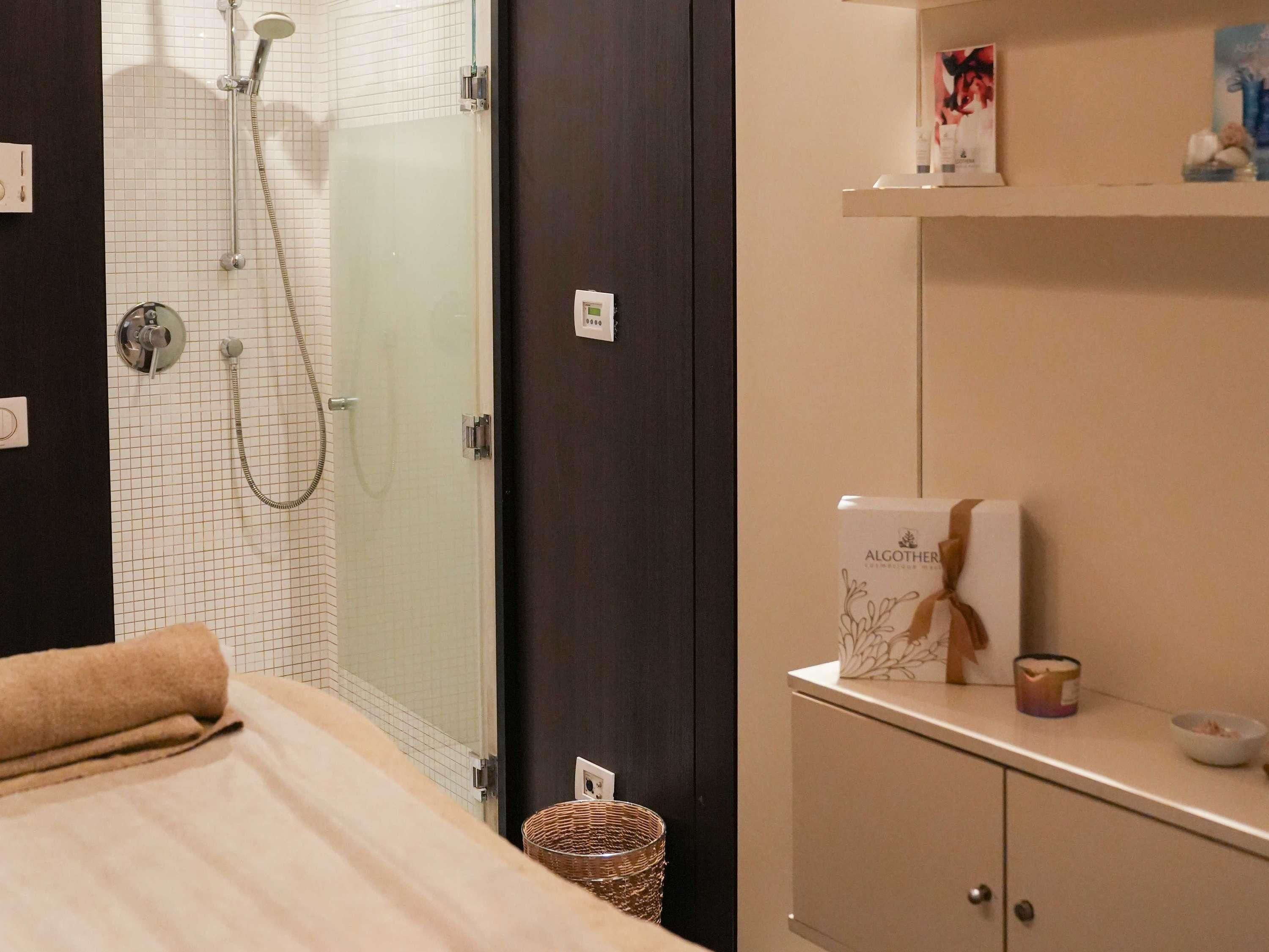 A bathroom with a glass shower enclosure, a towel on the floor, a cabinet with toiletries, and a wall-mounted light switch.