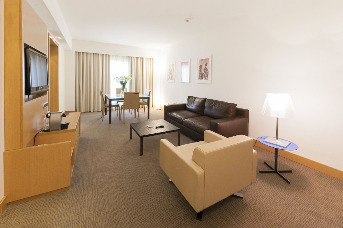 Habitación de hotel con sala de estar que incluye un sofá, una mesa de centro, un televisor, una mesa de comedor con sillas y una lámpara.
