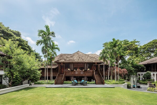A traditional wooden building with a thatched roof, surrounded by lush greenery and palm trees. - 137 Pillars House (Chiang Mai)