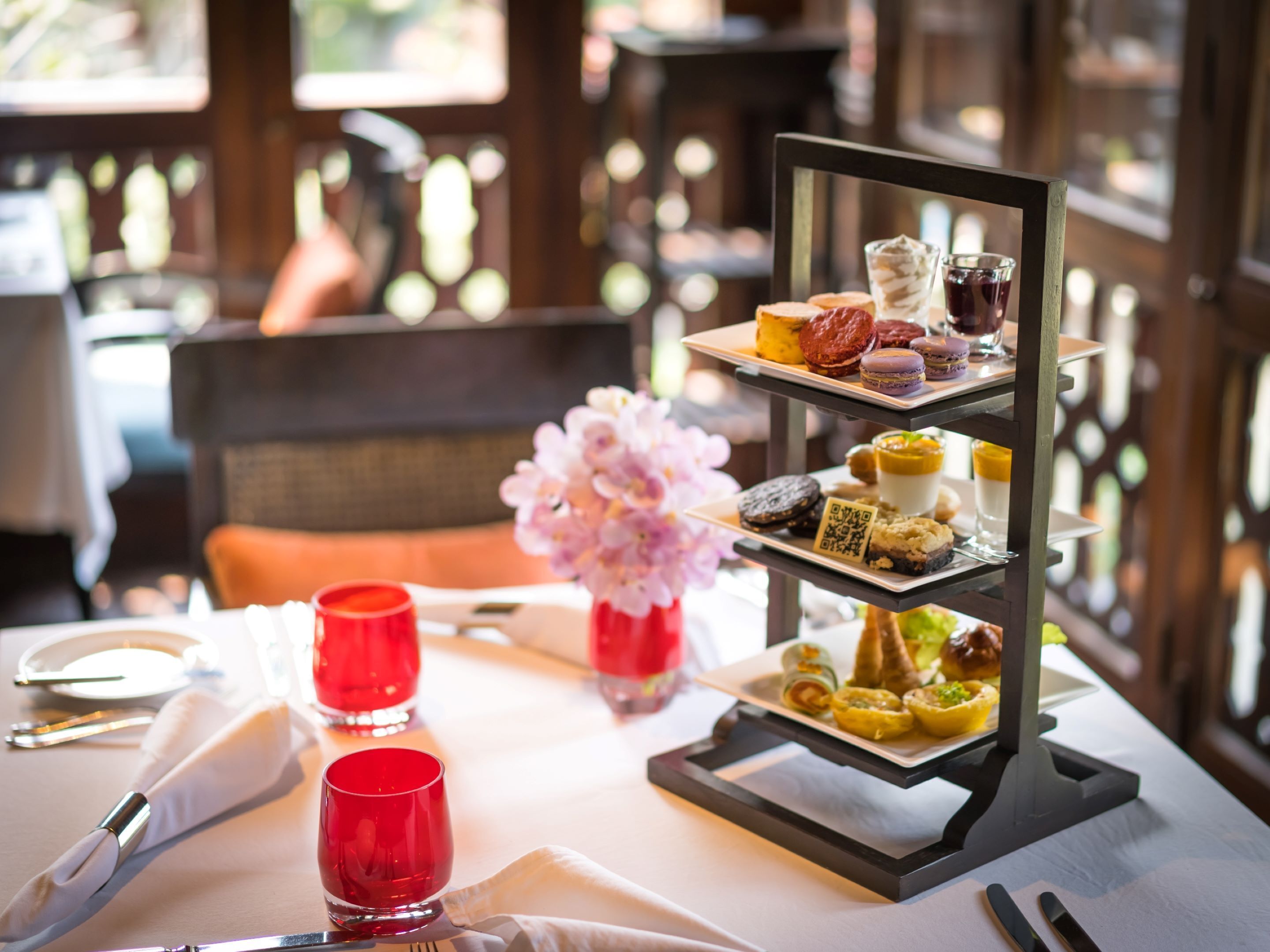 Un pastelero con postres variados y bebidas, un jarrón con flores, y una mesa dispuesta con un vaso rojo y una servilleta blanca.