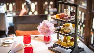Un pastelero con postres variados y bebidas, un jarrón con flores, y una mesa dispuesta con un vaso rojo y una servilleta blanca.