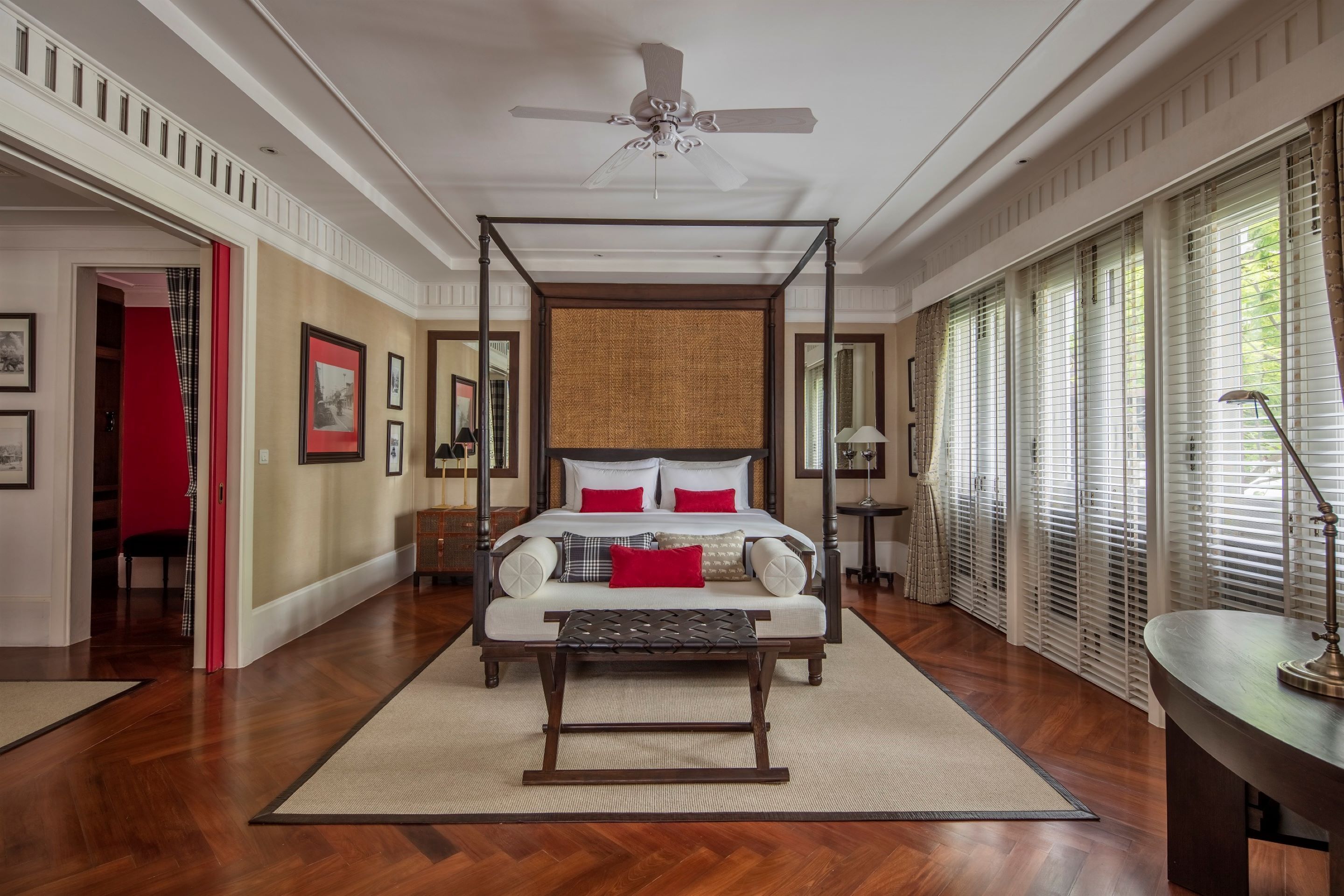 A spacious bedroom with a four-poster bed, a ceiling fan, and large windows with blinds.