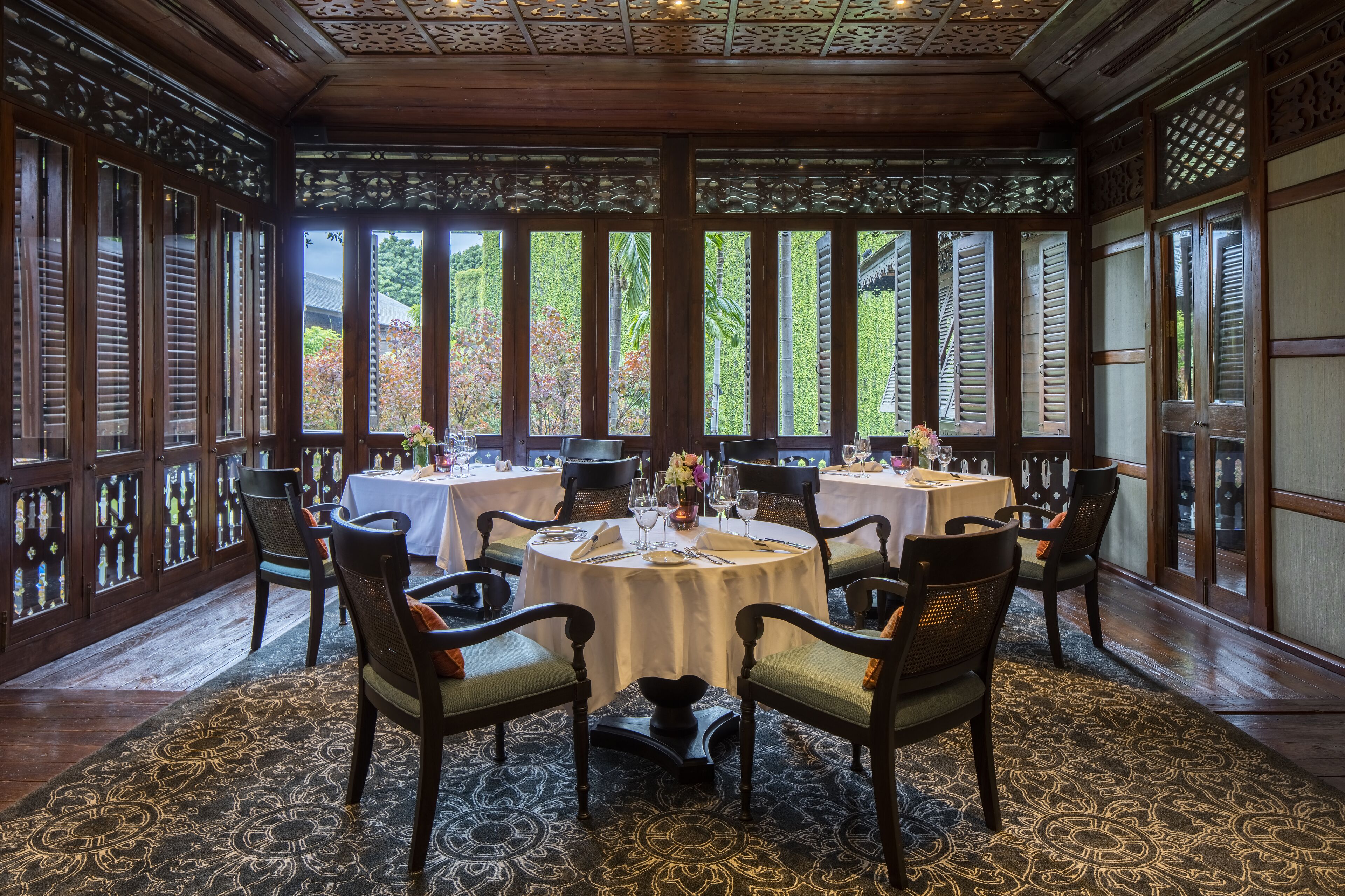 A traditional dining area with wooden furniture, patterned carpets, and large windows with decorative glass panes.