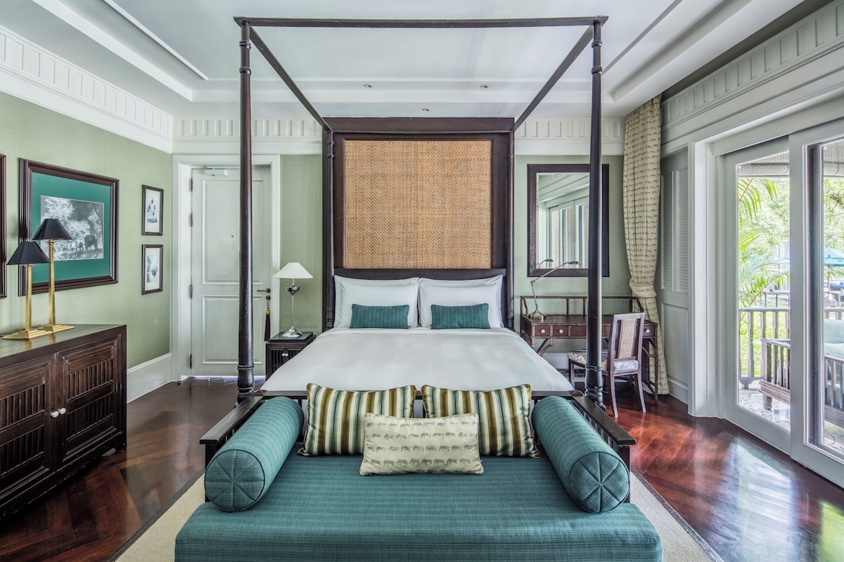 A four-poster bed with a canopy, a sofa with striped cushions, a wooden dresser, and a large window with a view of greenery.
