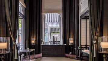 A room with a large window, dark curtains, a small table, a cabinet, and two lamps.