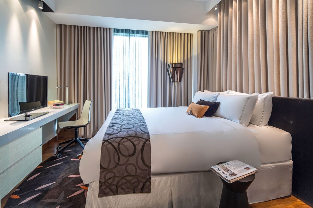 A hotel room with a bed, a desk with a computer, a chair, a lamp, and curtains.