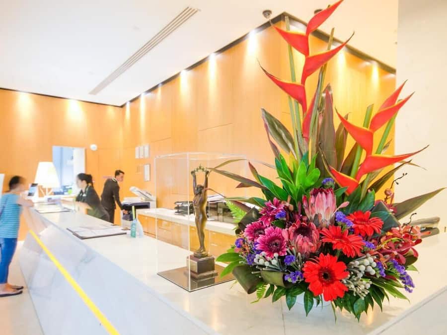 A reception desk with a floral arrangement, a statue, and a counter with a person standing behind it.